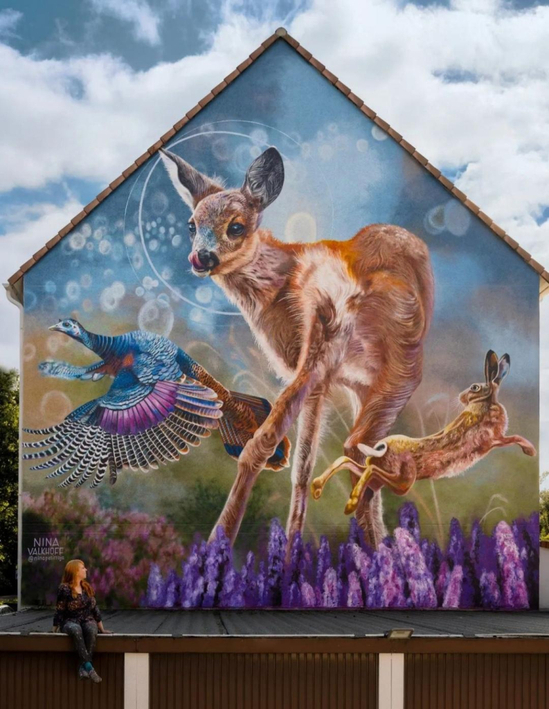 Streetartwall. A beautiful and imaginative mural featuring a light brown deer, a blue and purple turkey bird and a brown hare was spray painted on the outside wall of a two-storey house. The little fawn stands in a lilac meadow between a leaping rabbit on the right and a colorful, flying turkey bird on the left. The background is blurred and shows a green meadow and a blue sky. The deer's head is marked with thin light circles. The artist is sitting on the canopy in front of her mural. The picture was drawn and designed based on photos of Colinda van Binnendijk. It deliberately looks a little kitschy, but has a serious intention.
Info: The artist wanted to draw attention to three animals living in Germany that may be hunted: 
"This mural is a silent protest against hunting for pleasure. By depicting these animals being free and fluffy, I hope to make people in Gelsenkirchen happy, and whoever knows the meaning behind this painting will hopefully start to appreciate alive animals more than 'unalive' ones."