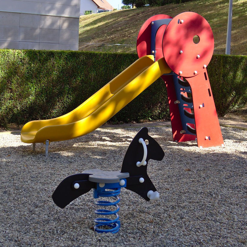 2 jeux pour enfants : un cheval (noir) sur ressort (bleu) au 1er plan, un toboggan rouge, bleu et jaune au 2nd, sur une zone mi-ombragée. Une haie derrière sépare d'un bâtiment, et d'une bute inclinée en fond.