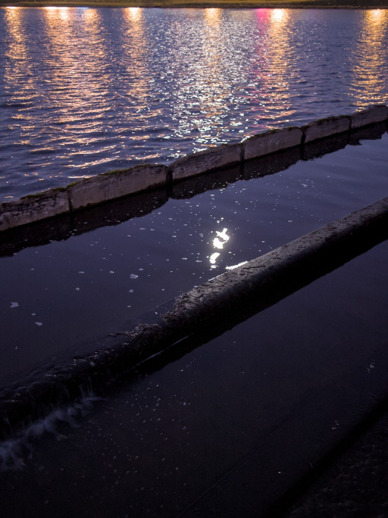 Photo de nuit : étendue d'eau , barrée en travers par un muret empêchant un courant trop important. L'eau derrière forme de petites vagues, devant est plus plate. Au 1er plan, un autre muret parallèle plus bas qui laisse l'eau s'écouler (trop plein). Se reflètent des lumières artificielles et la lune déformée.