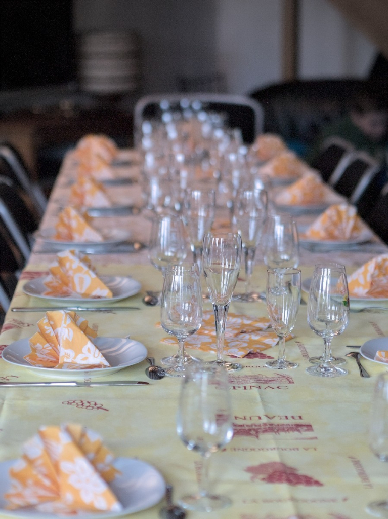 Une grande table dans une pièce éclairée par lumière naturelle côté gauche, vue depuis un des bouts. Elle comporte de nombreuses assiettes, verres à vin & flûtes à champagne. Les serviettes en papier sont orange et blanc, la nappe plutôt jaune avec des inscriptions de villes et des dessins de raisins.