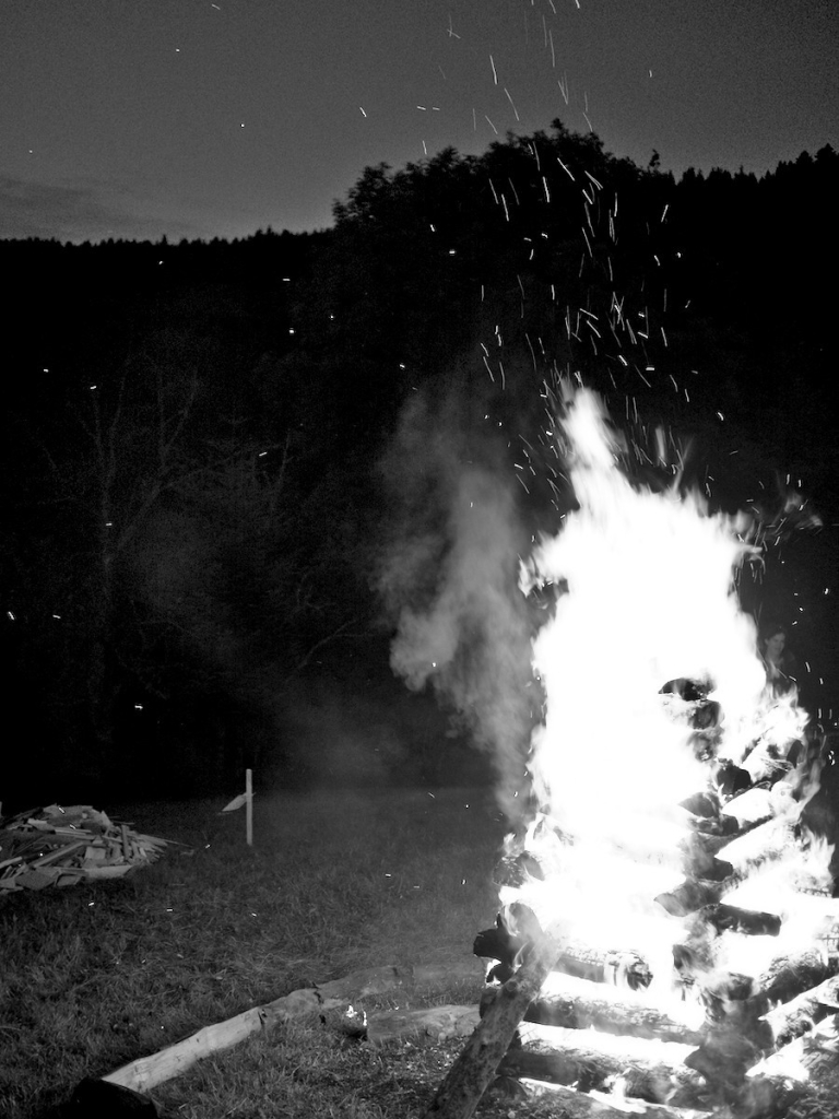 Photo en noir & blanc, avec au 1er plan sur la droite : un feu en "chavande", empilement ordonné de bois en forme de pyramide, tout autour de l'herbe et un tas de bois pour alimenter le feu à gauche en retrait. Un gros tiers supérieur de l'image est noire avec la silhouette d'une forêt en haut, surplombée par un ciel découvert de crépuscule.