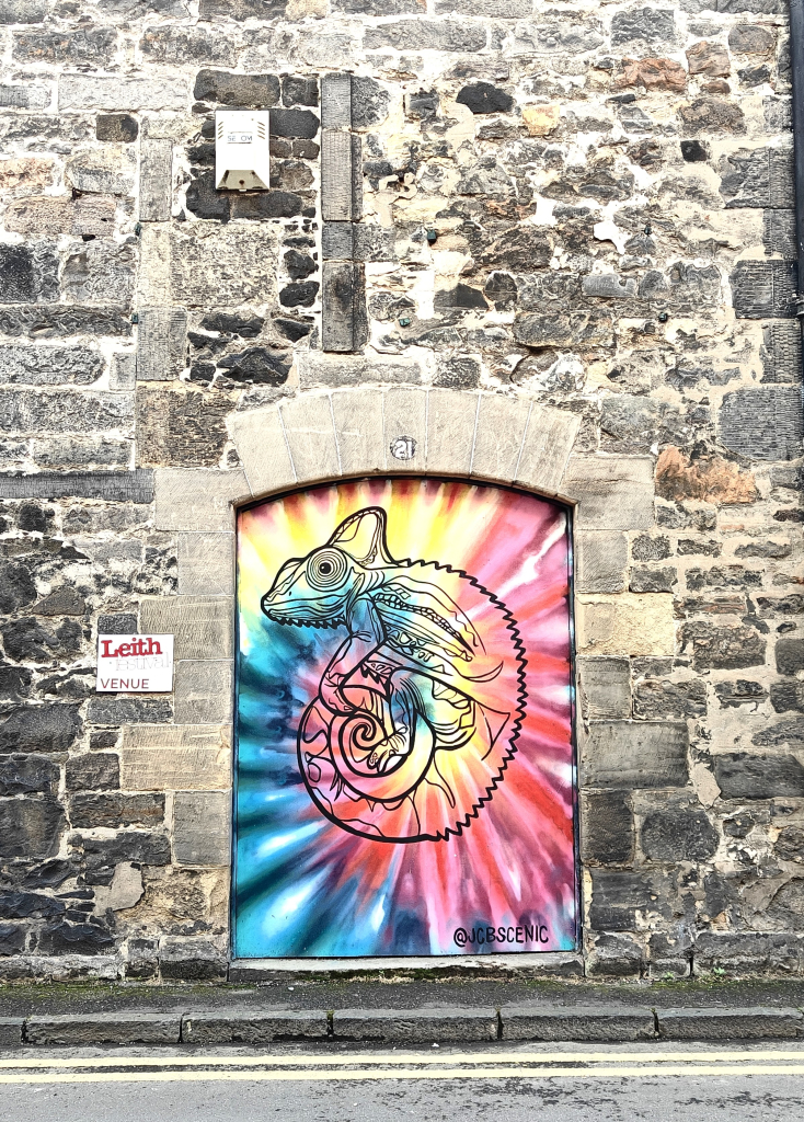 A colour photograph in portrait mode of a mural in Leith. The mural occupies the space of a former doorway into an old stone building, possibly a former warehouse. 

The mural shows a chameleon painted in black outline and the space filled with yellow, red and blue paints, creating a rather psychedelic image.