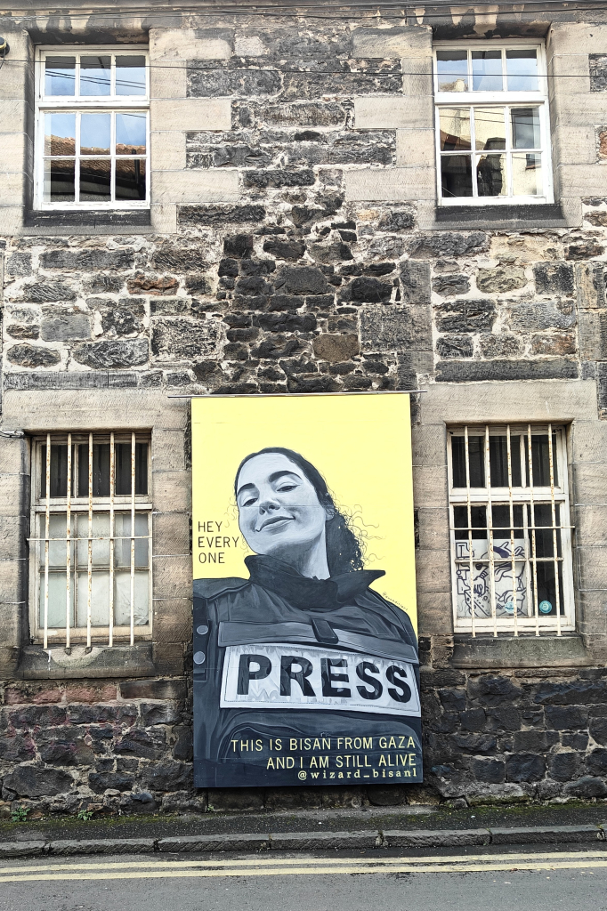 A colour photograph of a mural on a residential stone building, possibly a repurposed warehouse, in Leith. Between two windows, the height of one storey, the mural is the portrait of a young woman showing her head and torso. The woman is wearing a flak jacket bearing the word "PRESS" across her chest. She is smiling.

The background in the mural is a vibrant lemon-yellow. To the left of her head is the text "HEY EVERY ONE" and at the bottom of them mural is the text "THIS IS BISAN FROM GAZA AND I'M STILL ALIVE" with the social media tag @wizard_bisan1.

I sincerely hope the text is still true and that she is alive. (The Instagram account bearing her name still seems to be active.)