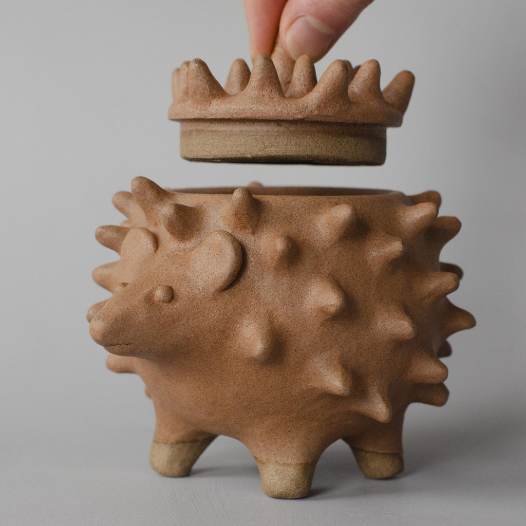 neutral grey background with a sculpture of a brown hedgehog on four legs with a lid that is being lifted by two fingers.  