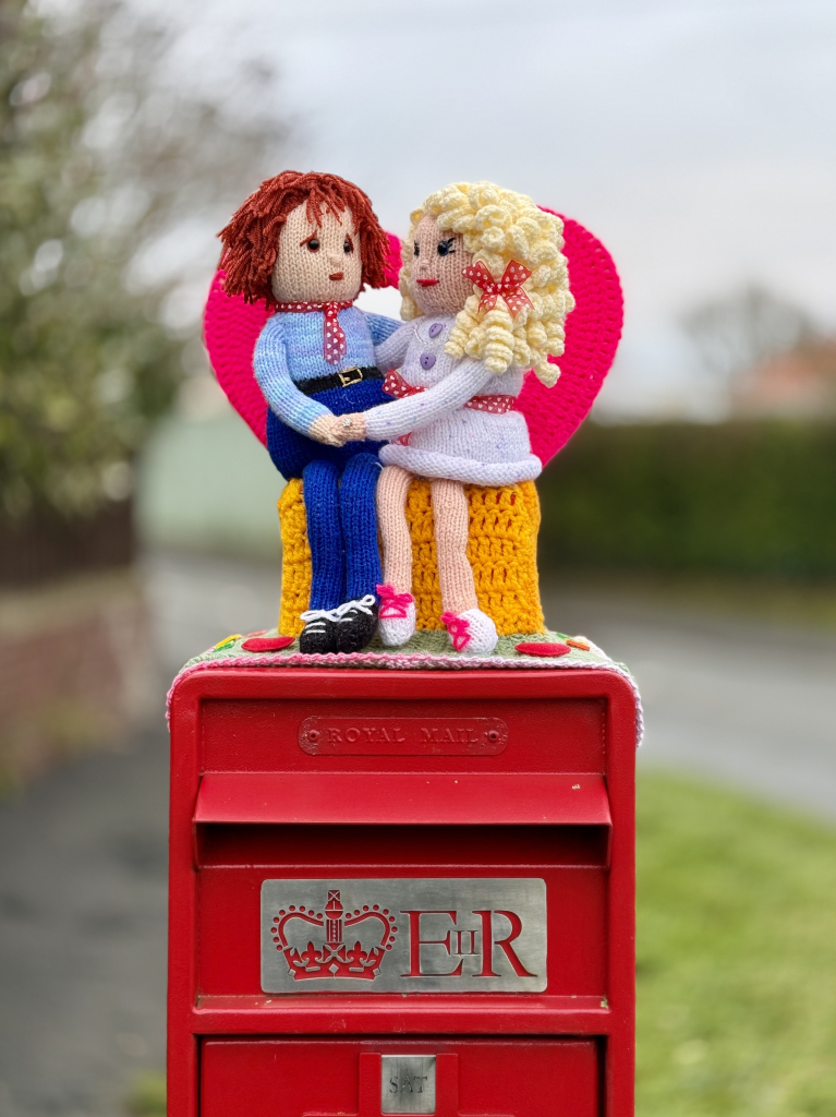 Red postbox with knitter figures of two people sharing an embrace while sat on a love seat. They look very much in love ❤️