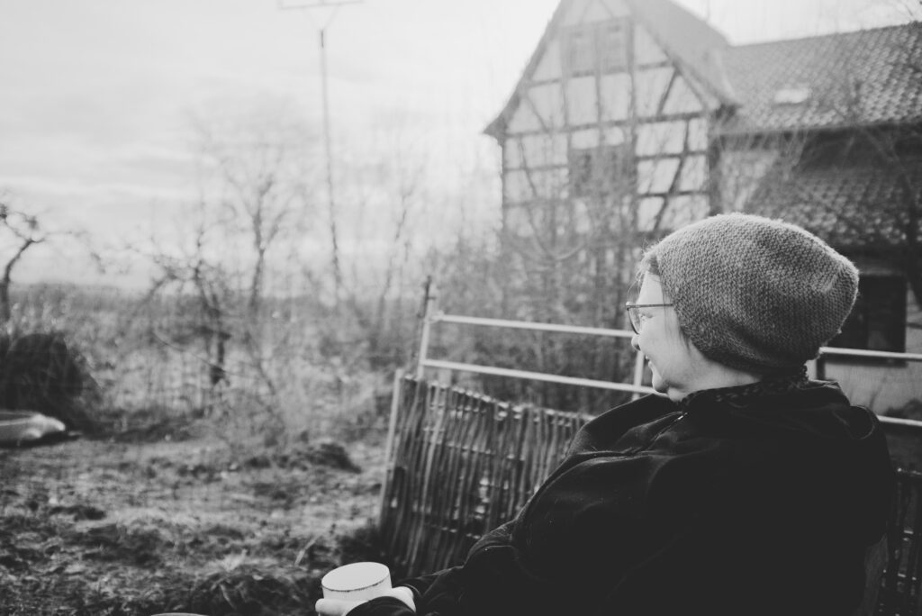 A woman sitting in a garden in front of a cottage with a cup in her hands.