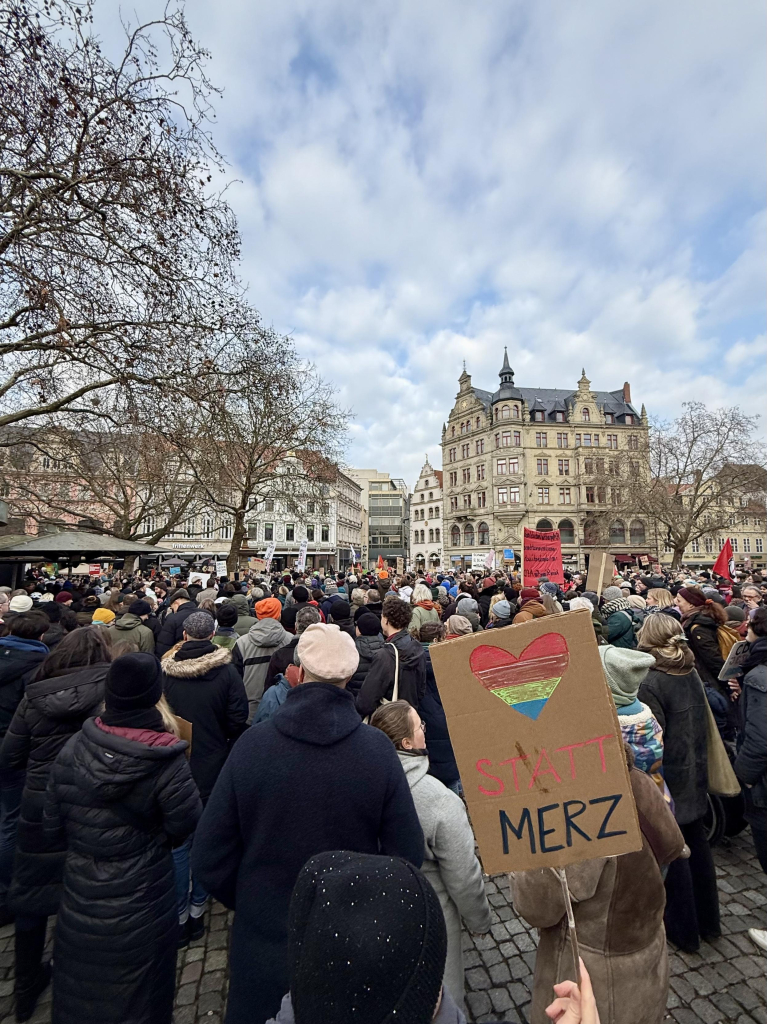 Eine große Menschenmenge versammelt sich auf einem öffentlichen Platz und hält Schilder während einer Demonstration. Die Szene zeigt historische Gebäude, kahle Bäume und einen bewölkten Himmel. Ein prominentes Schild zeigt ein Herz mit Regenbogenfarben und den Text "❤️STATT MERZ".