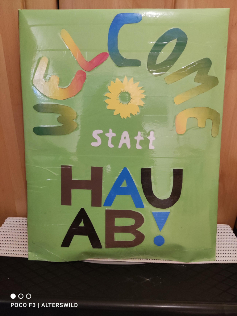 Demoplakat: hellgrüner Untergrund, darauf im Halbkreis "WELCOME" in Regenbogenfarben. Im Halbkreis eine gelbe Sonnenblume und in weißer Schrift: "statt"
Darunter in Großbuchstaben
HAU
AB!
Buchstaben in kackbraun, blau und schwarz.