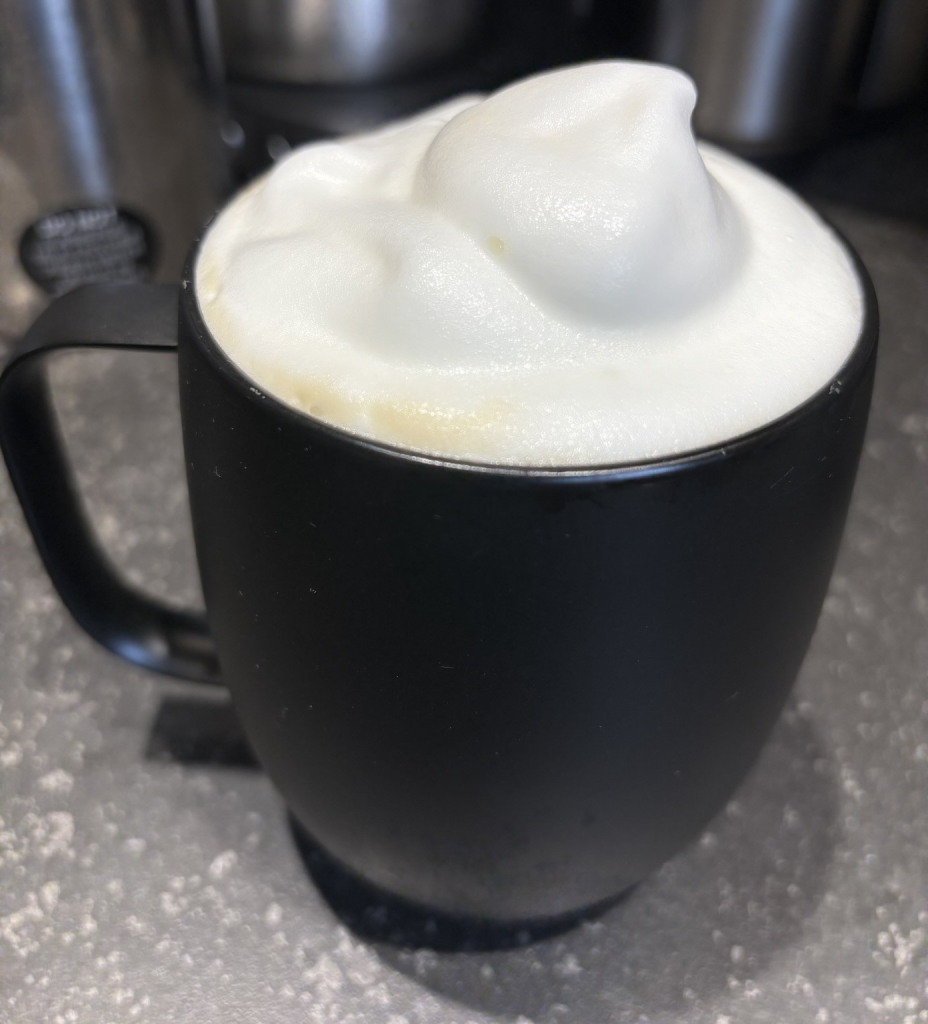 A black mug filled with frothy white foam, likely from a coffee or cappuccino, sits on a textured surface.