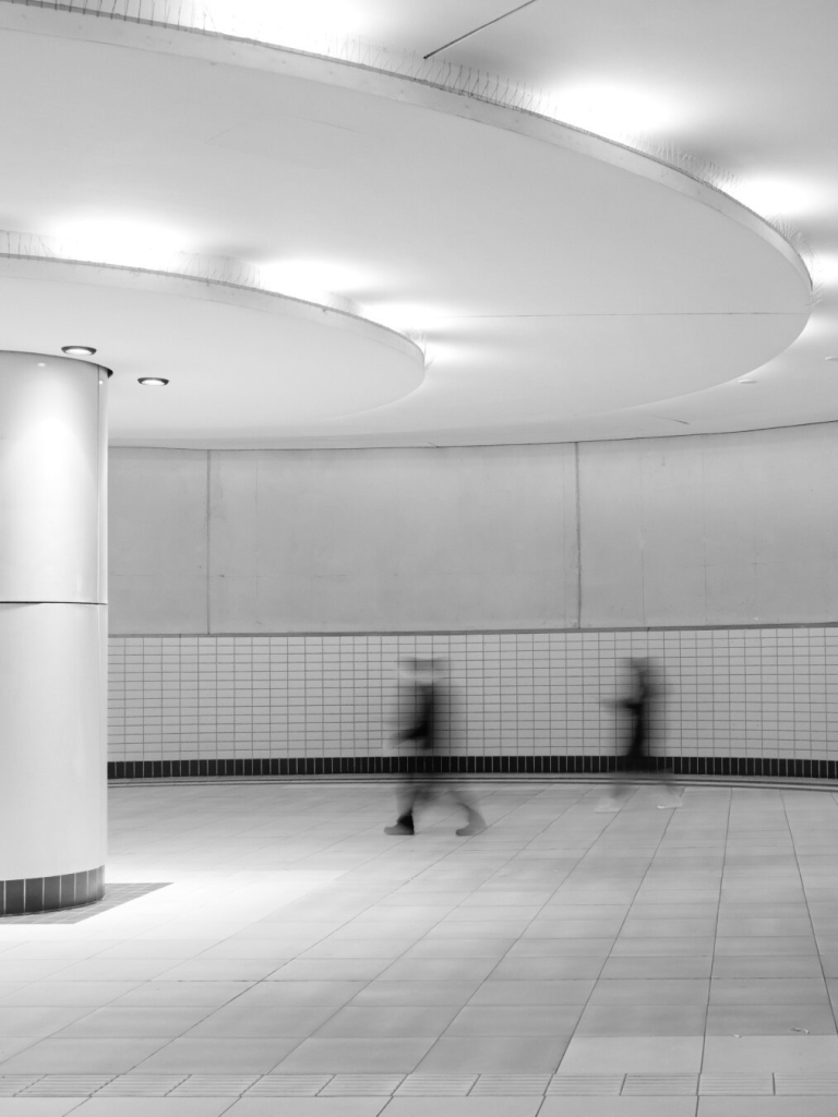 two pedestrians dressed in black hurry through the passageway of a bright white tiled subway station. the silhouettes are blurred by the long exposure time and thus appear ghostly.