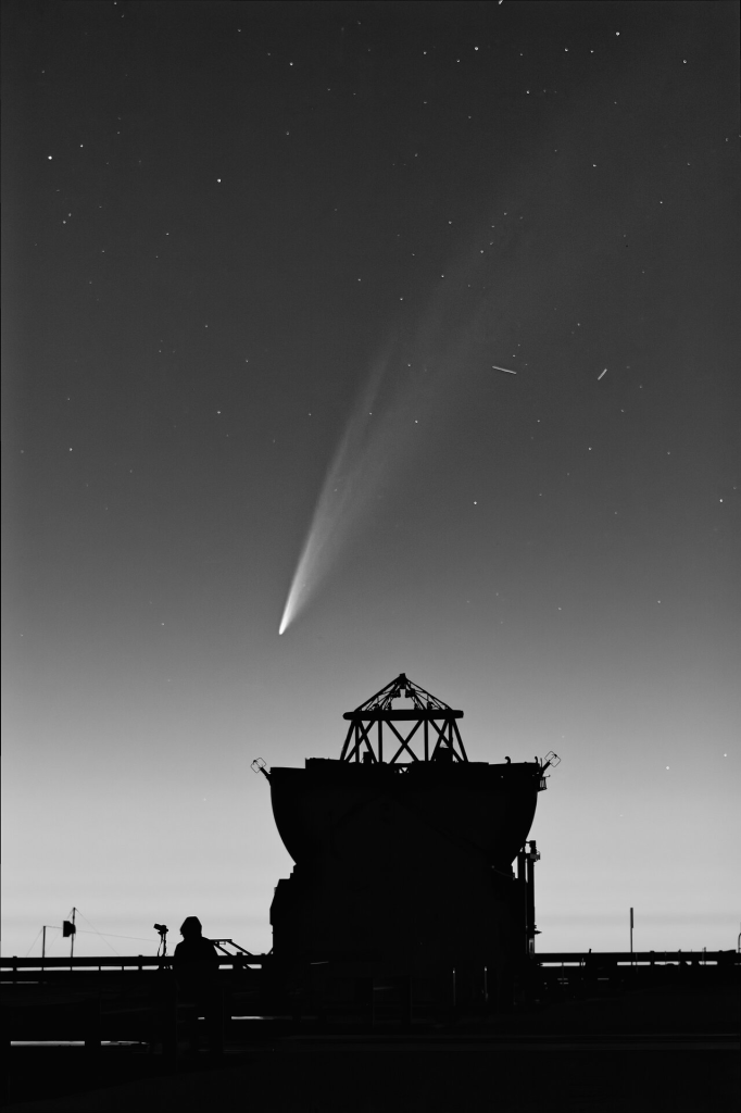 This black and white picture shows a small telescope and a large portion of the night sky. At the forefront, the silhouette of a small telescope reveals its small dome and central structure. Besides it stands a man, wearing a hood. The background of the picture shows the night sky, although only a few stars are visible, as well as a few short white strikes which indicate moving objects. The ’star’ of this composition, though, is the comet C/2024 G3 (ATLAS), which covers most of the frame and looks like a giant feather in the sky (or a bushy cat’s tail). The core of the comet (its tip) is bright white, while its tail becomes more diffuse as you go away from the core.