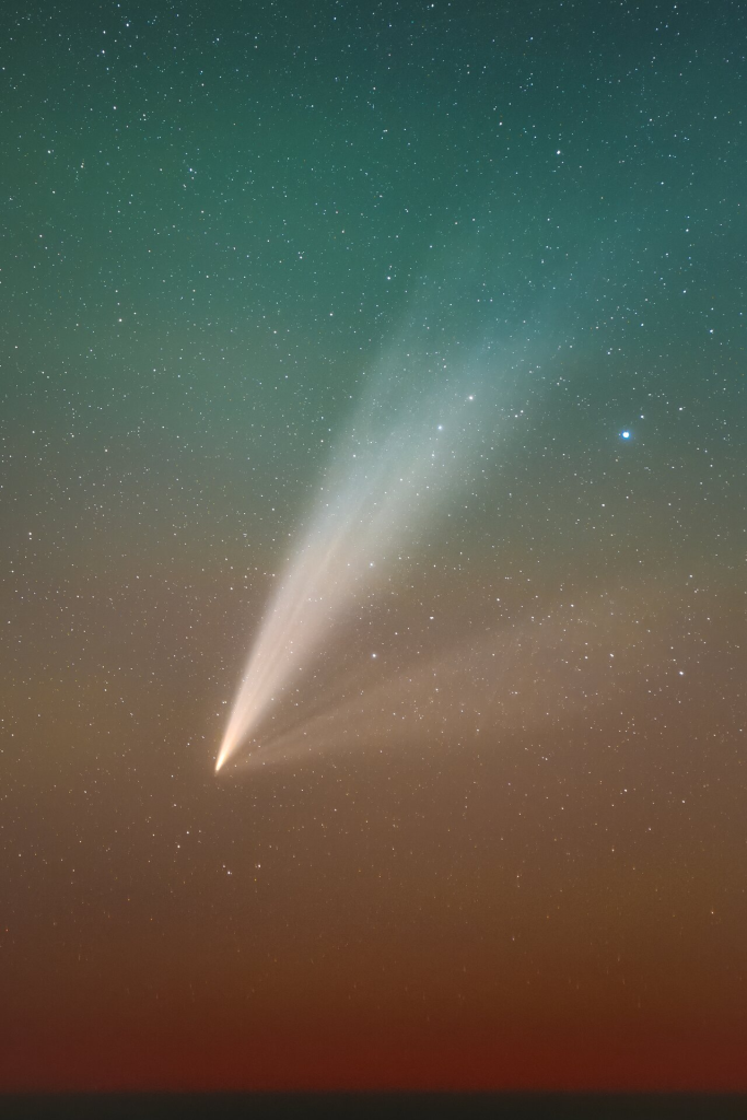 In a gradient from orange-red to green-blue, this image captures the Comet C/2024 G3 around dusk. The background is covered by countless stars of all sizes. At the forefront, the comet appears as a white feather-shaped object. The core of the comet (its tip) is bright white. From there, two tails originate: towards the right projects a fainter tail, almost like a veil. Towards the top projects a brighter tail, which looks almost like the bushy tail of a cat and which becomes fainter the further you go from the comet’s core.