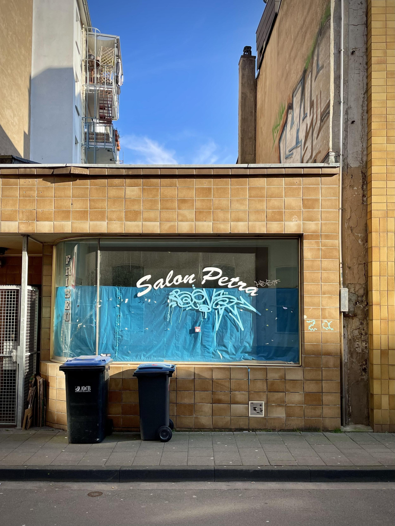 das bild im hochformat zeigt einen sonnenbeschienenen flachbau mit schaufenster, auf dem in geschwungener schrift „salon petra“ steht.
das gebäude ist bräunlich-ockerfarben gefliest. das schaufenster ist von innen mit blauer folie abgeklebt. unter „salon petra“ ist ein hellblauer graffiti-tag zu sehen. vor dem haus stehen zwei mülltonnen. links und rechts sind hohe häuser wahrnehmbar. dazwischen und über dem flachbau ist blauer himmel mit leichter bewölkung zu sehen.