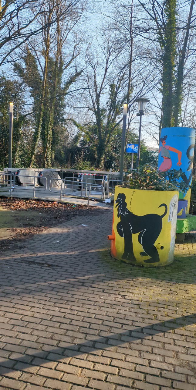 Picture of entrance to "Delta" subway station close to the ULB Fosdem conference location.  It's beginning February, trees mostly have no leeves, and there are wall paintings: a kind of animal/human in black on yellow ground, as well as a yellow/blue towerlike structure with some red and black graffiti.  A stairway and two machine-rolling stairways are visible, one of them currently blocked.  