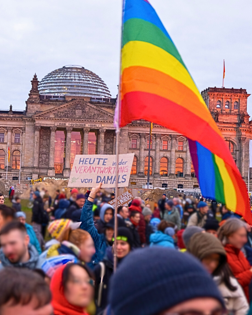 Foto einer Demonstration vor dem Reichstagsgebäude, in der Bildmitte eine Regenbogenfahne.