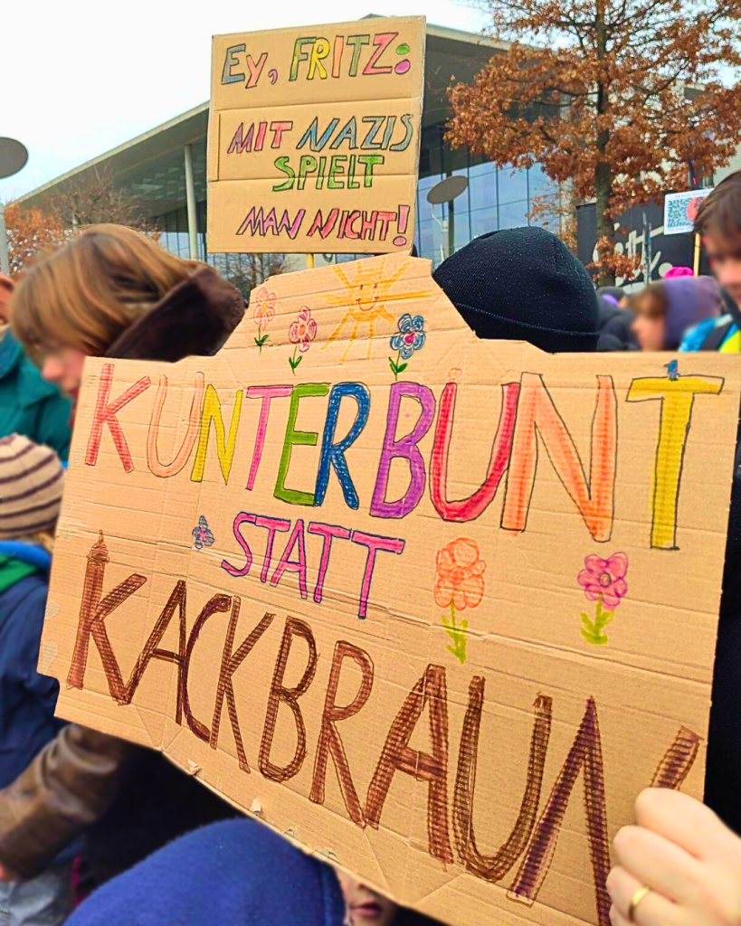 Zwei Schilder auf einer Deminastration vor dem Reichstagsgebäude, beide mit bunten Wachsmalstiften auf Pappe gemalt. Auf einem der Text: "Ey, Fritz: Mit Nazis spielt man nicht!", auf dem anderen: "Kunterbunt statt Kackbraun."