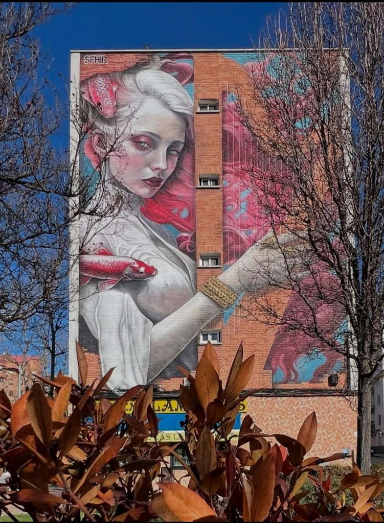 Streetartwall. A mural with the image of a mystical female creature playing a lyre was sprayed/painted on the exterior wall of a four-storey modern building. In the middle of the wall is a row of windows. The Mural shows a modern version of a siren from the Greek legend of Orpheus. The graceful lady has a pale but young face, white hair that turns pink in length and blows in the wind. She wears a tight white dress, a golden bracelet and stands turned to one side. She plays an upright lyre and is accompanied by pink fish swimming around her. She looks determined and proud. 
Info: In Greek mythology, the sirens lured sailors with their hypnotic song in order to kill them. The Greek hero Orpheus fought the sirens' song with his lyre and turned them to stone. The mural continues the story fictitiously. The siren returns, stronger than ever, having taken over Orpheus' powers and gazing majestically at passers-by.