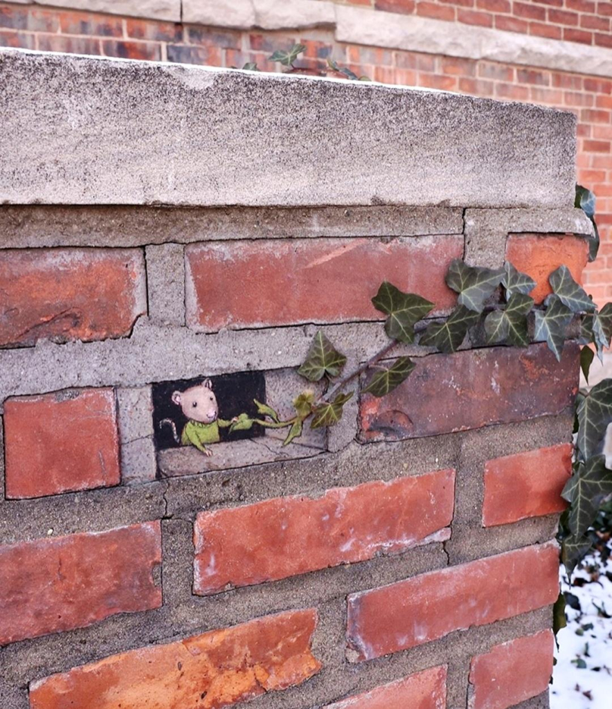 Streetart. A little mouse (called Emile) and a brick-sized hole have been drawn in chalk on a red brick wall. Emile, the mouse, wears a green woolen sweater and looks out of the hole. She reaches for a real green plant tendril growing towards the wall. The lower leaf in her hand looks like a little heart. Title: "Emile's new friend is a little clingy, but evergreen."