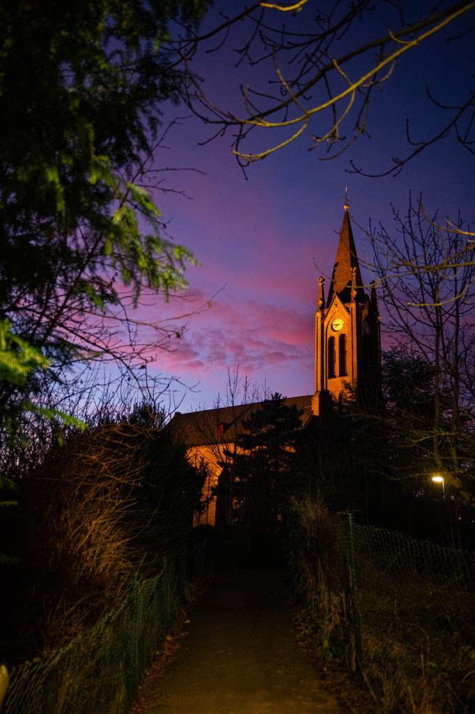 Das Foto zeigt eine angeleuchtete Kirche, im Hintergrund blauer Himmel der mit roten Wolken des nahenden Sonnenaufgangs bedeckt ist.
Unten sieht man einen kleinen Weg der zu der Kirche hinauf führt. Rechts, links und oben sind Sträucher und Äste zu sehen und rahmen das Bild ein. 