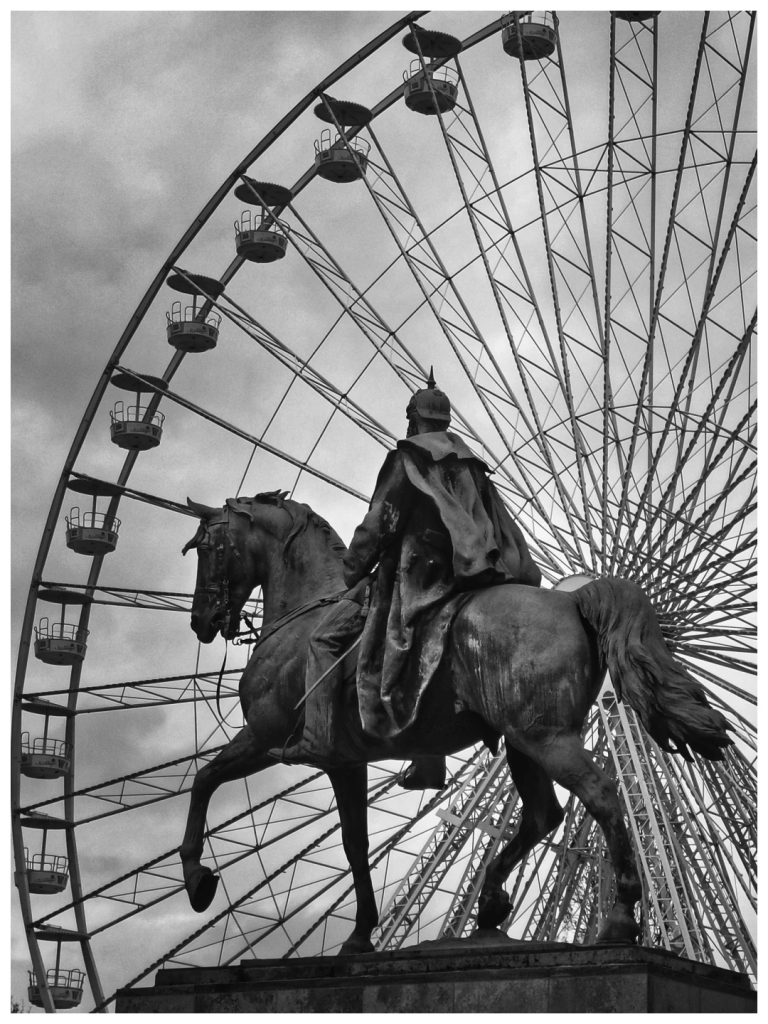 Das Schwarzweissfoto im Hochformat zeigt im Vordergrund ein bronzenes Reiterstandbild, das Kaiser Wilhelm I. als Feldherrn in Generalsuniform mit Mantel und Pickelhaube darstellt. Im Hintergrund ein Teil eines 50 m hohen Riesenrades mit offenen Gondeln, das Foto nahezu ausfüllend.