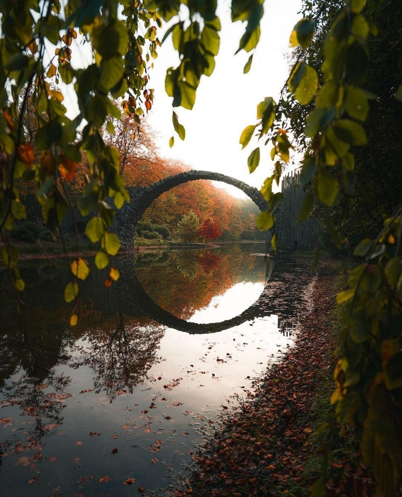 Photography.  A color photo of an old arched bridge over a lake in an autumnal landscape. The bridge is bent upwards and, together with the reflection in the water, forms a circle. The sun's rays falling from above change the lighting conditions in this circle. All around are autumnal deciduous trees on the bank. A beautiful nature shot and the photo has a wonderful atmosphere
Info: The 19.80-meter cantilever arch bridge was built by a landowner beginning in 1866 as part of a garden ensemble and is also known as the Devil's Bridge. The bridge was featured in the Hollywood film "Matrix Resurrections" and the 2017 German film "Der Zauberlehrling".
