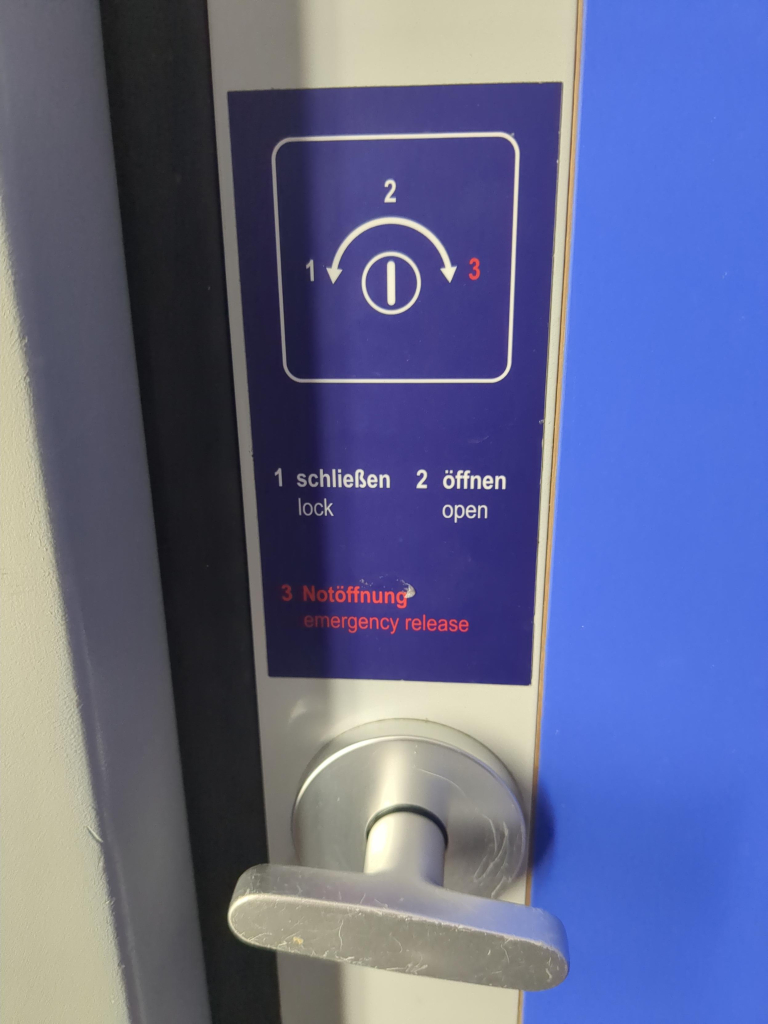 Photo lock/release and sign as described in more detail in main post. The sign is in blue with white text, except the emergency release text which is red. The diagram positions are numbered, and the numbers have a key in German and English. The twister is T-shaped, rounded and aluminium.