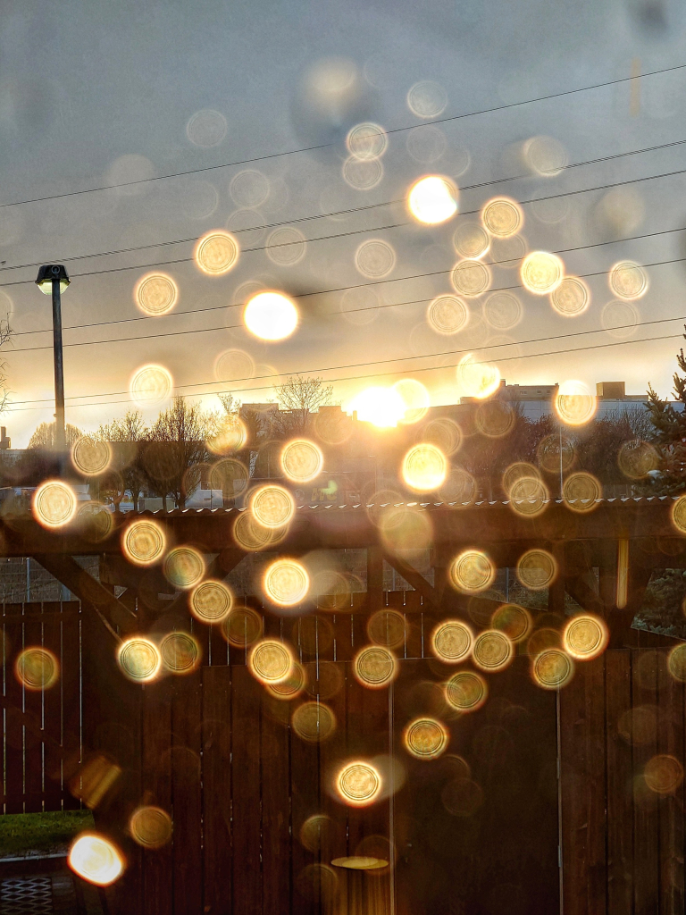 Blick durch ein Fenster von innen nach außen. An der Außenseite der Scheibe haften Regentropfen, die im Bild verschwimmen und durch den Lichteinfall der Sonne als Lichtpunkte erscheinen. Im Hintergrund ist die Bebauung eines Dorfes zu sehen.