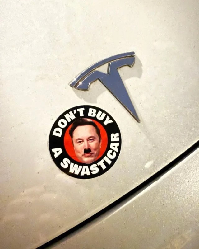 Round sticker on a Tesla with a picture of Elno with a Hiltler-esque moustache & text “Don’t buy a swasticar”