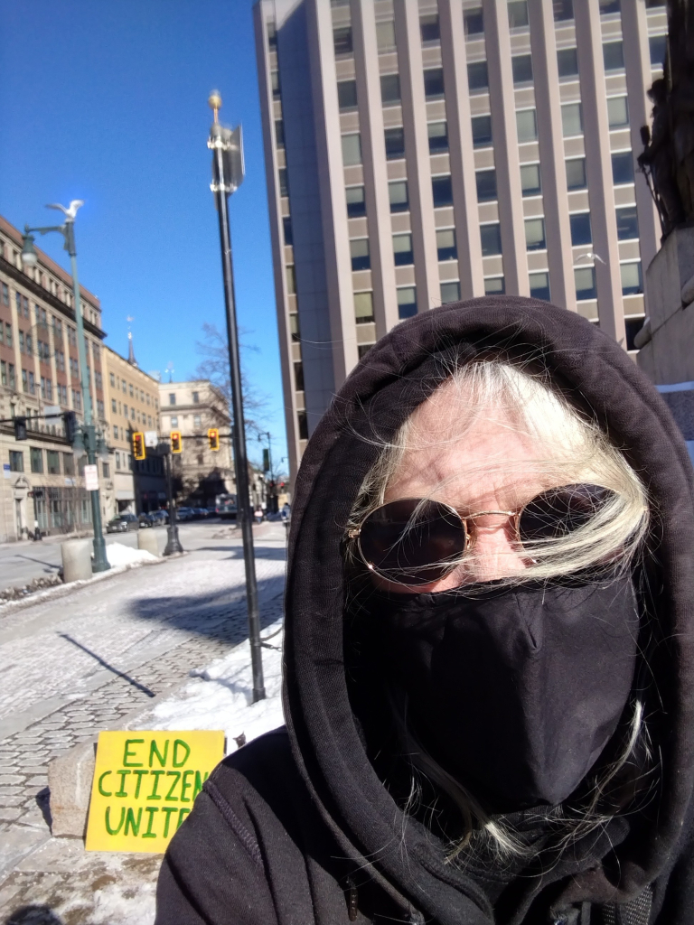 A person with white hair, wearing sunglasses, a black mask, and a black hoodie, standing on a street corner in the city. A sign that reads, "End Citizens United" is behind them.