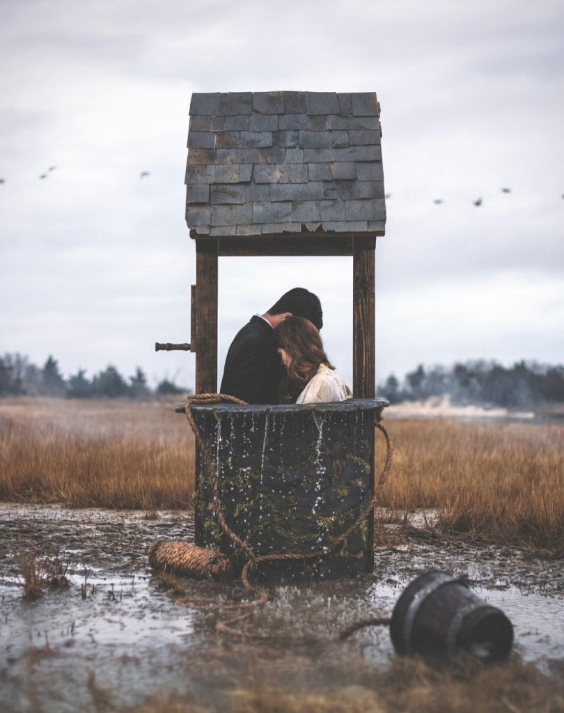 Photography and Art. A color photograph of a man and a woman in a covered fountain. The surreal photo is a composition of photo and self-made sculptures. The picture shows a foggy gray landscape with a field of grain in which an old stone well with a roof stands. The well is surrounded by water and a wooden bucket lies in a puddle in front of it. Two people are standing in the well, holding each other tightly. You can see the upper bodies of the couple, a young man in dark clothing, while the brown-haired woman is wearing light-colored clothing. Water flows from the fountain. A few birds fly high into the gray sky, as if they want to escape the dreary place. The overall mood of the picture is mysterious, characterized by loneliness and silence.
Info: Nicolas Bruno, an artist who transforms his dreams and nightmares into surreal imagery. His photography is also a way of regaining control of the narrative of his sleep paralysis experiences. 
PhotoInfo: Nikon D810, Nikon 50mm
Digital Photography and self-made sculptures 