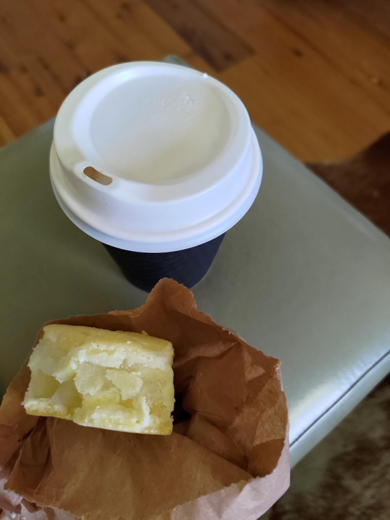 View from above of a take away coffee cup and an apple slice in a paper bag on a small green stool on a timber floor with brown rug