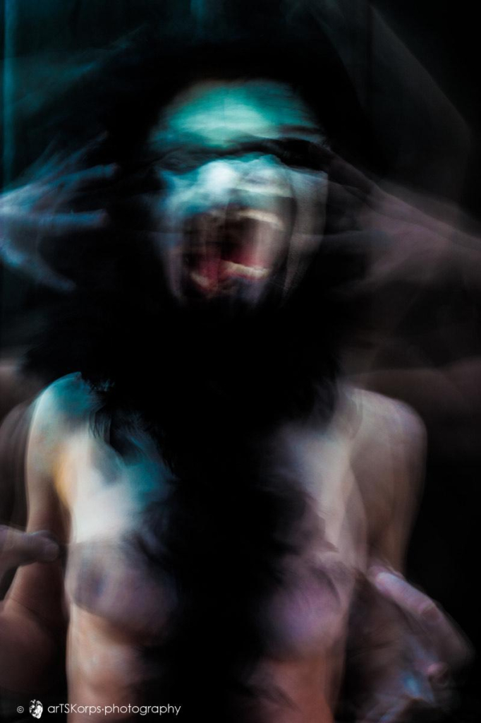 Colour photo in portrait format of a naked female torse in blurred motion. The person is crying with her mouth wide open, her hands with spread fingers to the sides of her head. Dominating colours against a black background are blueish greens