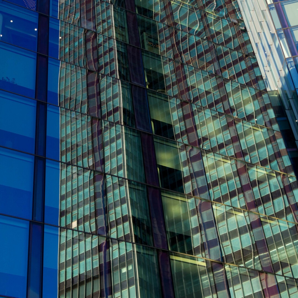 Photo of a wall of glass glossily reflecting a greenly towering building, two grids of tall windows with a shadowy recess between. Through the wall, divided into rectangles by thin black lines, light blinds are pulled down to varying levels.