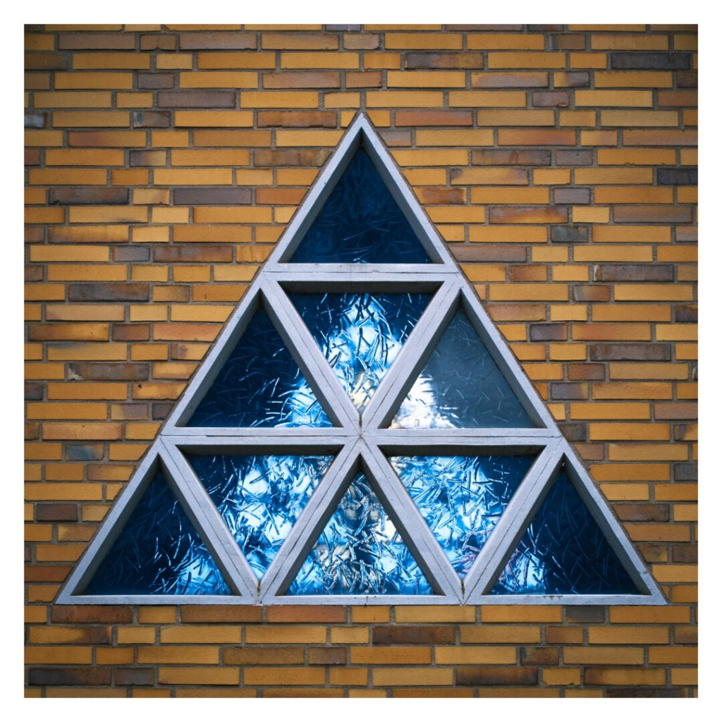 This square color photo, framed by a simple white border, features a triangular window set into a yellow brick wall. The window is composed of multiple smaller triangular panes of textured blue glass, which reflect light in a bright, almost crystalline way. Through these panes, another window is faintly visible, adding a subtle layer of depth. The warm brick background contrasts sharply with the cool tones of the glass.