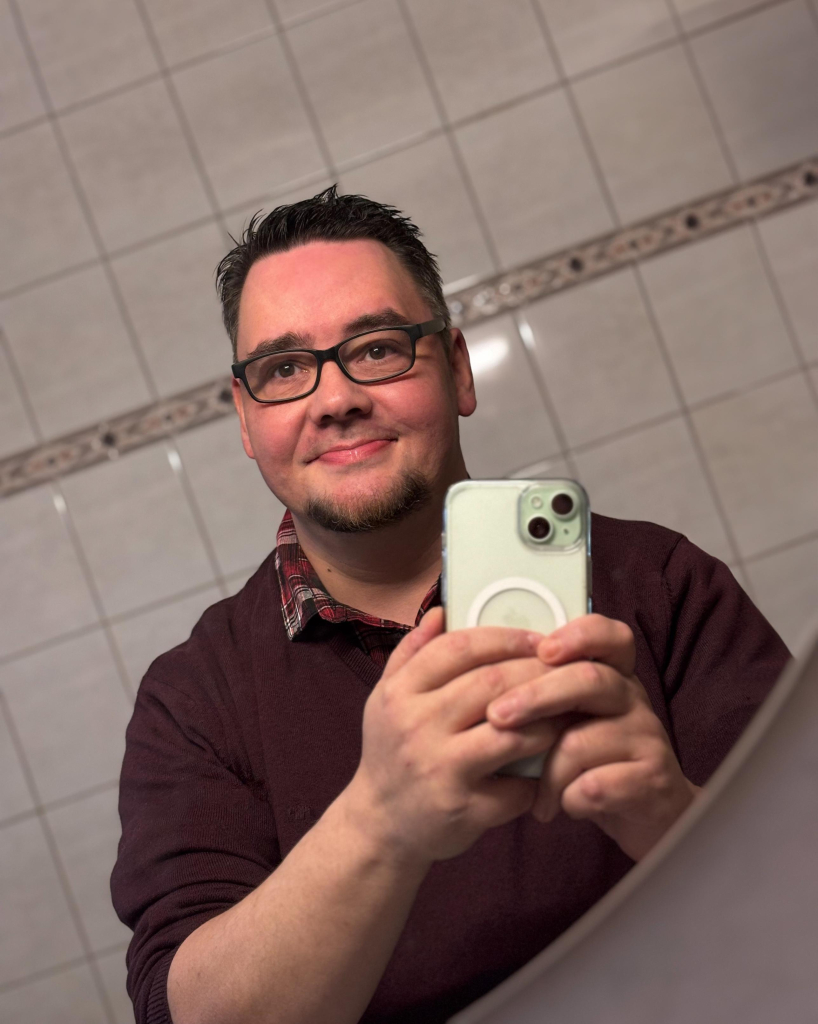 Motiv: Eine Person hält ein Smartphone und macht ein Spiegel-Selfie.

Bildformat: Hochformat.

Genre: Porträtfotografie, Selbstporträt.

Hintergrund: Geflieste Badezimmerwand mit einer dekorativen Bordüre.

Farben: Neutrale und warme Töne, hauptsächlich Beige und Braun.

Lichtsituation: Künstliche Beleuchtung aus einer Innenraumquelle, weiches Licht mit leichten Schatten.