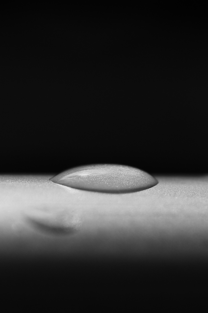 das schwarzweißfoto im hochformat zeigt eine starke nahaufnahme eines wassertropfens auf einer glatten, abgerundeten fläche.
der tropfen liegt flach auf einer grauen armlehne eines stuhles. die lehne ist als horizontaler streifen unterhalb der bildmitte zu sehen. die lehne hat eine sehr feine struktur bzw. musterung.
der flach aufliegende tropfen hat die form einer optischen linse. sie funktioniert tatsächlich auch wie eine linse. der tropfen gibt das bild der strukturierten armlehne wieder. sie ist ebenso wie die armlehne grau und ebenso texturiert.
der hintergrund ist schlicht schwarz. dadurch wirkt das bild minimalistisch.