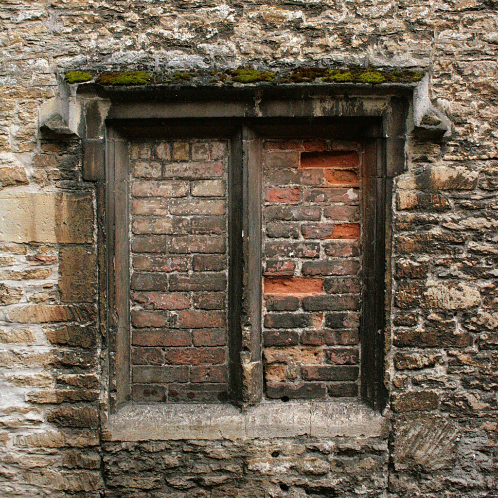 A Medieval window in a stone wall which has been bricked up with red bricks.