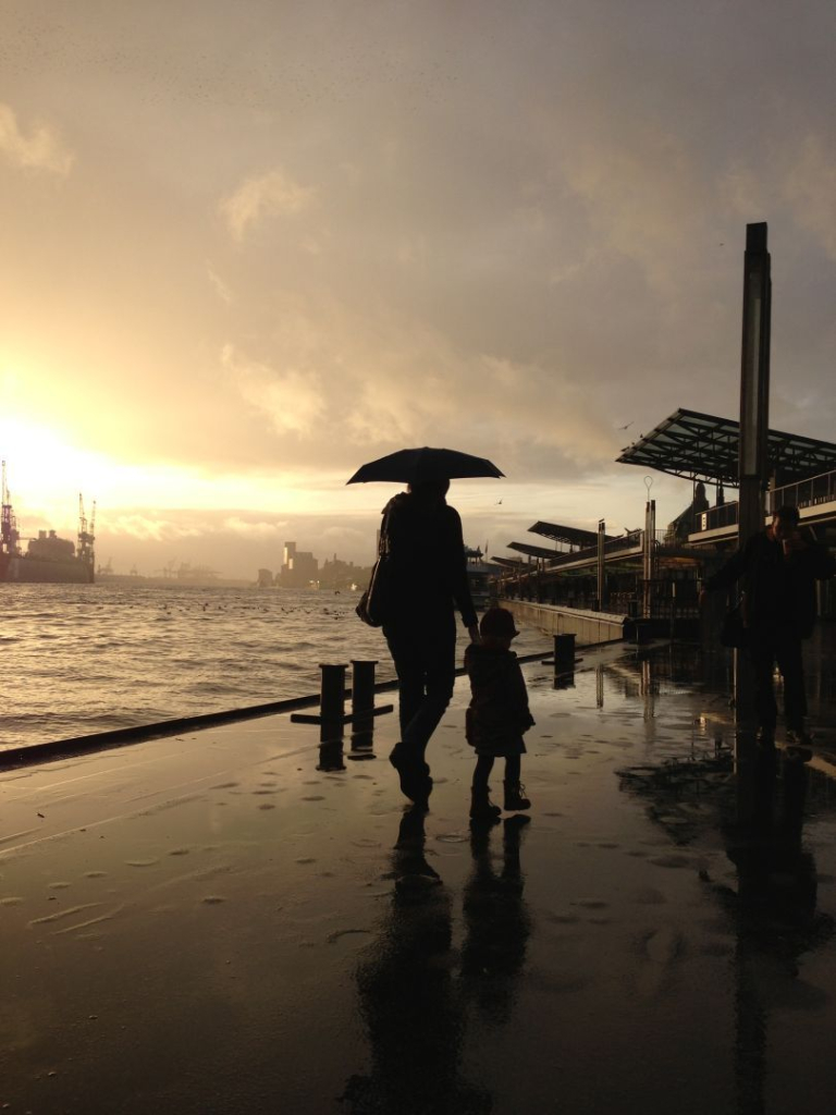 Ein regnerischer Nachmittag in Hamburg an den Landungsbrücken der Elbe. Eine Person mit Regenschirm geht mit einem Kind spazieren. Man erkennt nur ihre Silhouette und ihre Spiegelung auf dem regennassen Boden. Sie gehen der tiefstehenden Sonne entgegen, die dramatisch durch die Regenwolken bricht. Im Hintergrund erkennt man Docks und Gebäude.