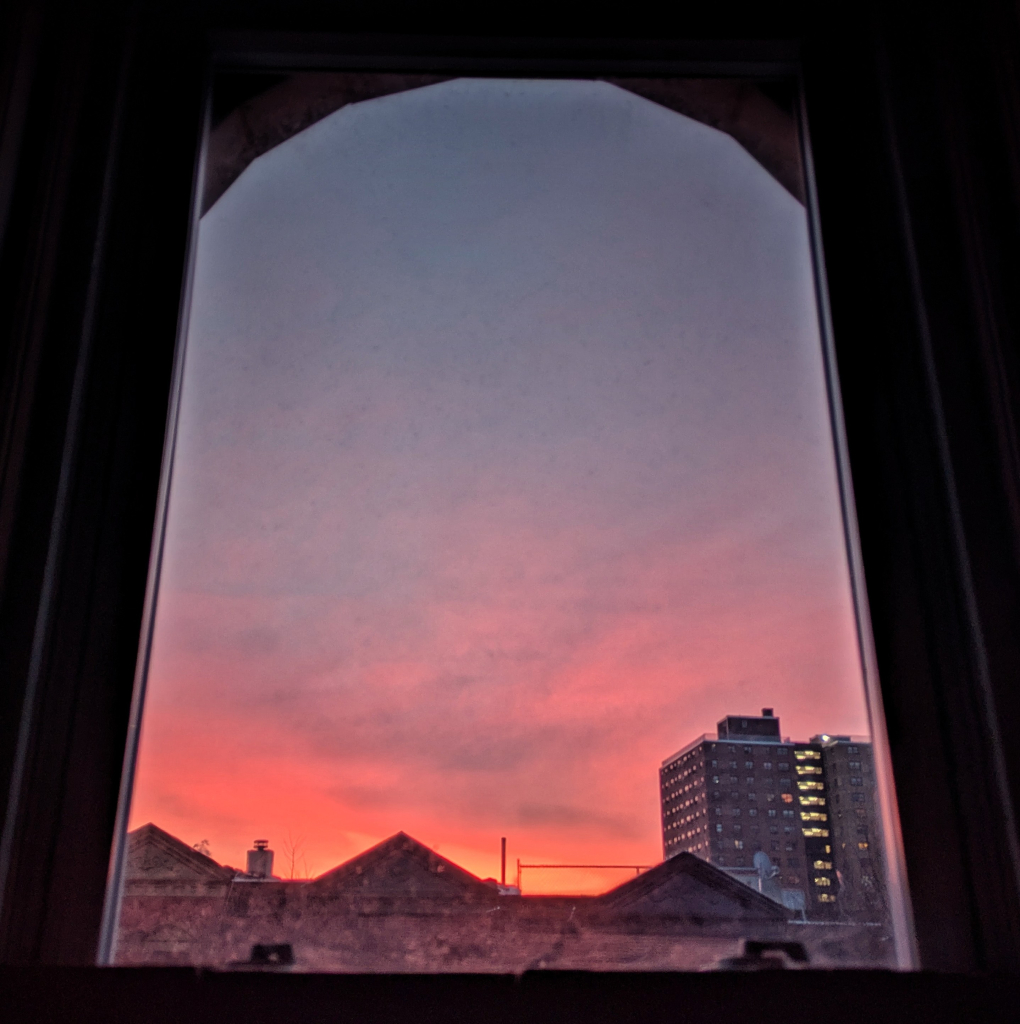 Looking through an arched window thirteen minutes before sunrise the sky fades from bright orange at the horizon to grey-blue up above. Pointed roofs of Harlem brownstones are silhouetted across the street, and a taller apartment building can be seen in the distance.