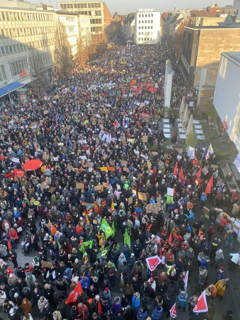 A large crowd gathered in an urban setting, holding various signs and flags during a protest. The scene is vibrant, with many participants wearing winter clothing. Buildings are visible in the background, indicating a public space being used for the demonstration.