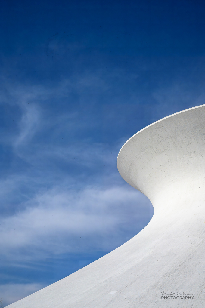 The curved form of a white concrete roof in a hyperboloid shape set against a blue sky with thin wispy white clouds. 