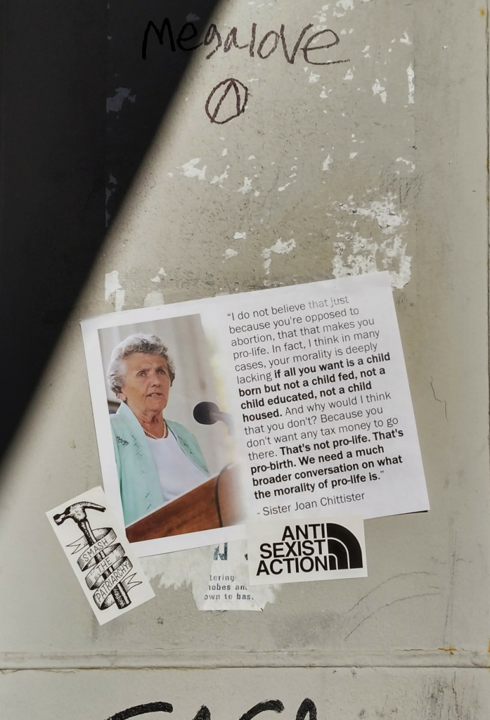 A flyer stuck to a wall. The flyer has an image of an older white woman with silver hair. Next to the image are the words:
"'I do not believe that just because you are opposed to abortion, that that makes you pro-life. In fact, I think in many cases, your morality is deeply lacking if all you want is a child born but not a child fed, a child educated, a child housed. And why would I think that you don't? Because you don't want any tax money to go there. That's not pro-life. That's pro-birth. We need a much broader conversation on what the morality of pro-life is."
Sister Joan Chittister

Holding the flyer in place are two stickers: One is a hammer with the words, "Smash The Patriarchy" wrapped around it on a ribbon. The other sticker has the words, "ANTI SEXIST ACTION".