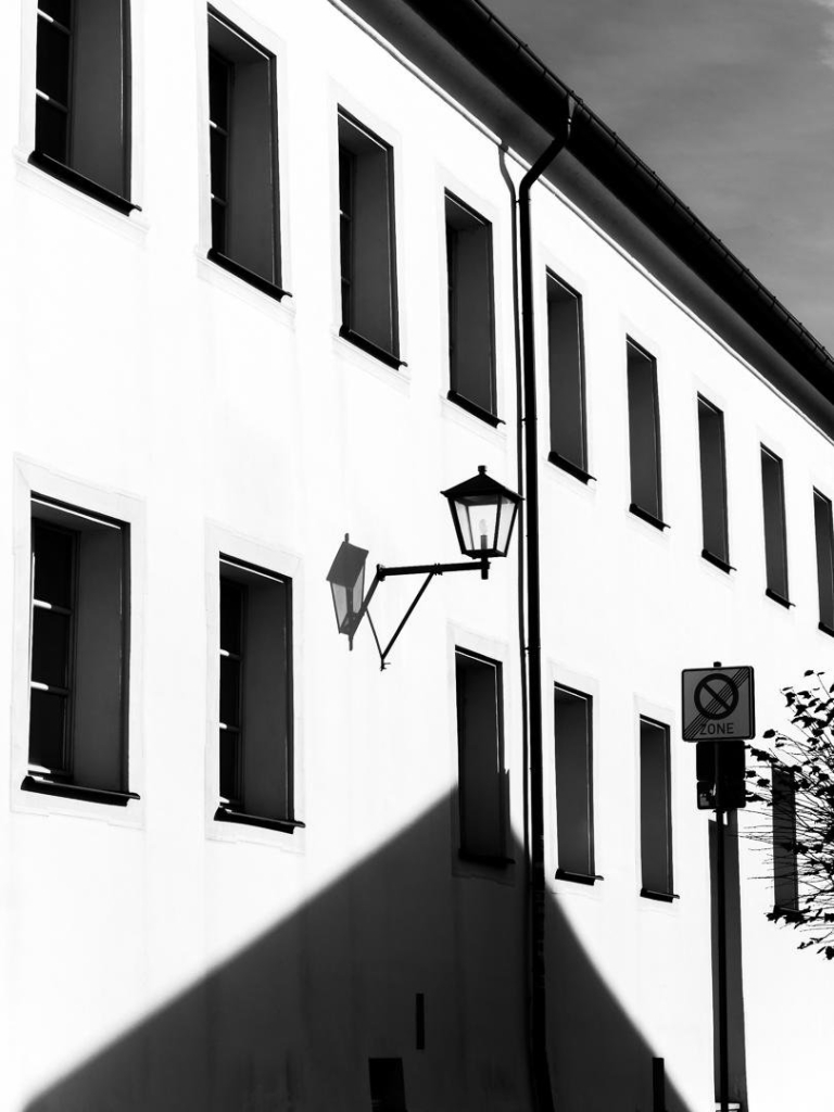 White wall with two rows of windows. A lamp hangs on the wall, in front of it are traffic signs. Hard midday light, black and white. Gutter, rain pipe create sharp shadows, from below a triangular shadow of a roof opposite.