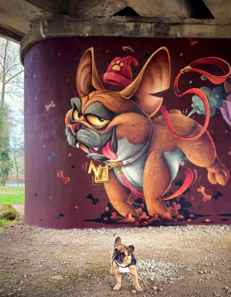 Streetartwall. A colorful comic mural with a cute bulldog was sprayed/painted on the concrete wall of a bridge pillar. The background is dark brown. The funny cartoon dog jumps happily in the air and wears a chain with the golden letter "N" for "Nestor" around his neck. His woolly hat is flying in the air over his head and his dog lead is twirling around. The photo also shows the "real" Nestor, a small French bulldog, standing in front of the wall. It is the artist's dog.