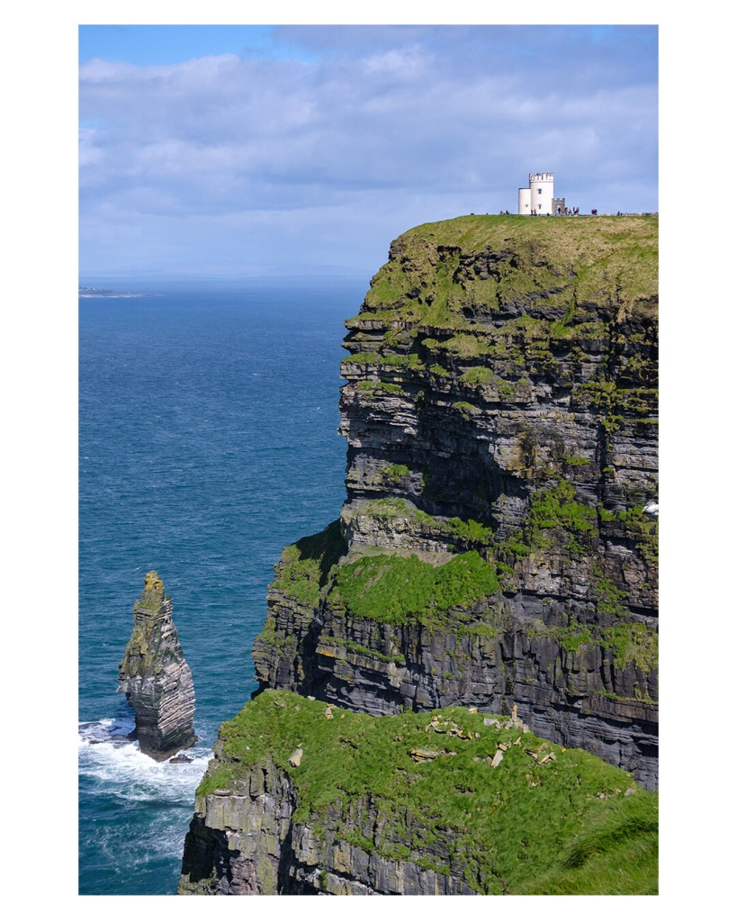 Foto im Hochformat. Blick auf ein riesiges Kliff am Meer. Links das steinige Kliff, es ist teils mit Gras bewachsen und man sieht unterschiedliche Schichten im Gestein. Links ist das Meer. Im Meer ist noch ein einzelner Felsen, der von der Brandung umspült wird. Rechts oben auf dem Kliff ist ein weißes Gebäude. Davor sind winzige Menschen auszumachen. Es verdeutlicht, wie hoch das Kliff tatsächlich ist. Der Himmel ist leicht bewölkt.