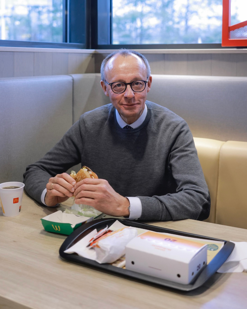 Friedrich Merz sitzt offenbar in einem McDonalds-Restaurant und hat einen Burger in der Hand. Darunter die offene Burger-Packung mit einem gut sichtbaren Logo. Daneben eine Getränkebecker ebenfalls mit Logo.