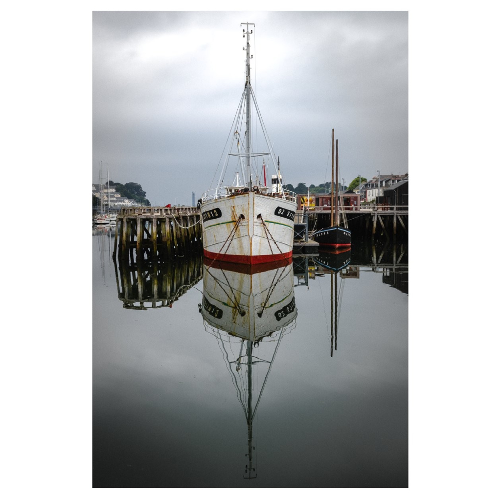 Blick auf einen Hafen der in einem natürlichen Meeresarm liegt. Wir sehen frontal auf den Bug eines Fischerboots. Das Boot spiegelt sich nahezu perfekt im spiegelglatten Wasser. Das Bild wirkt etwas düster, dank des grauen Wetters.