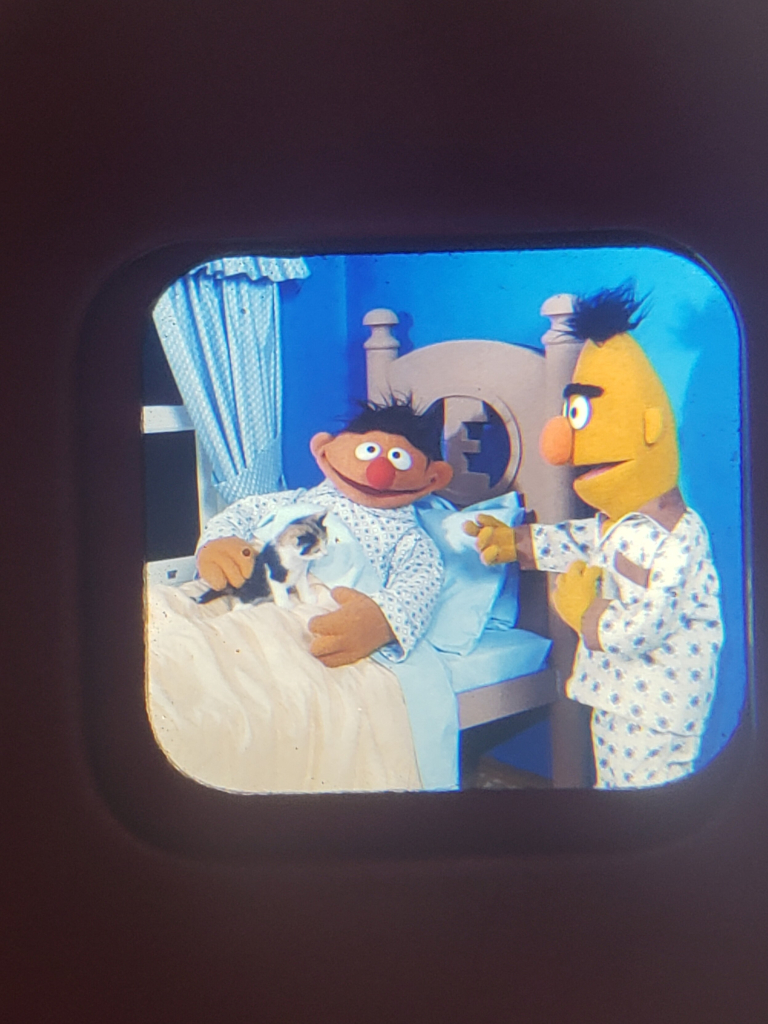 Bert, Ernie and the kitten get ready for bed. The kitten sits on Ernie's lap, Ernie tucked in, and Bert, also in pyjamas, stands nearby.