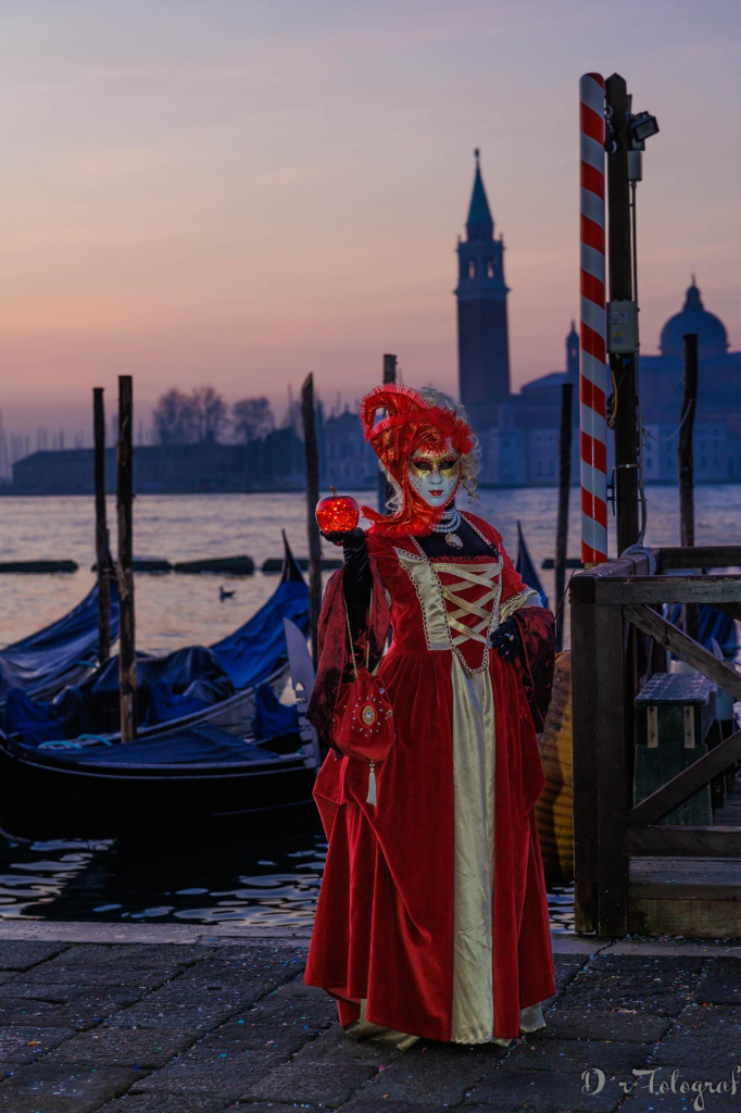 Venzianische Maske in Rot mit rotem Apfel in der Hand. Stehend vor Gondeln in Venedig in der Morgendämmerung. Im Hintergrund