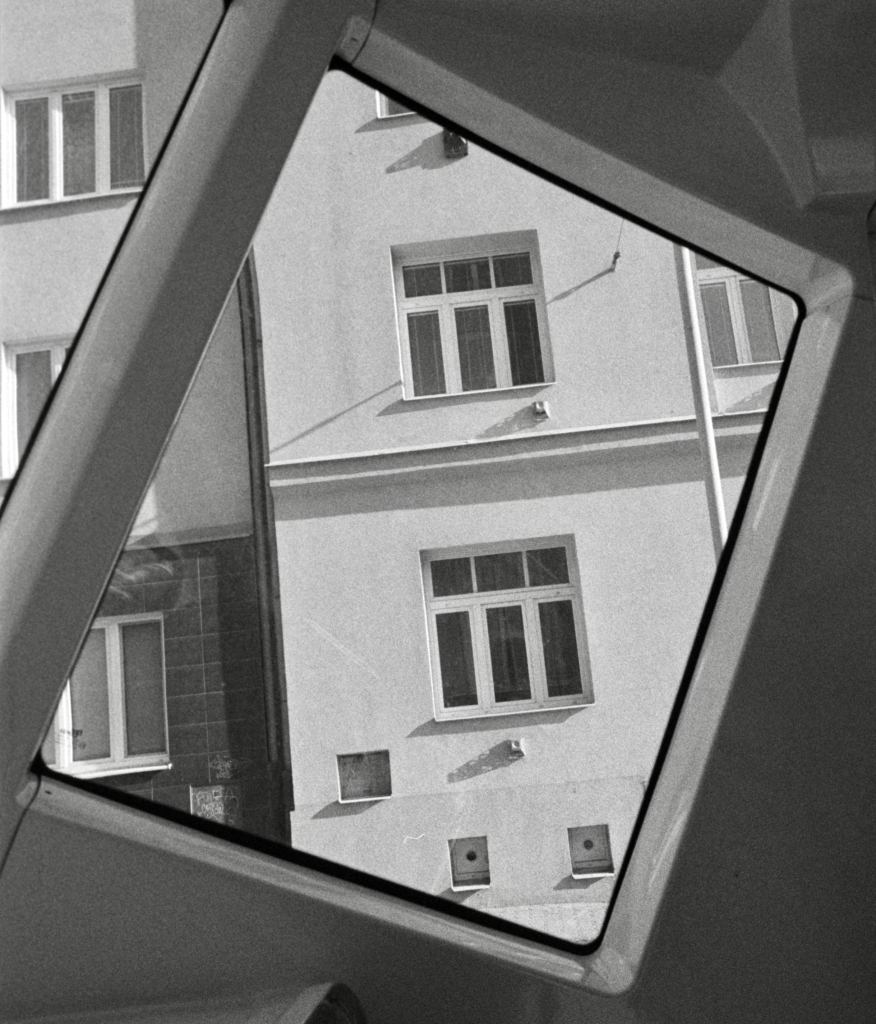 Black and white film photo. View from a tram through a diamond-shaped window. Behind the window, you can see the wall of a house with two windows